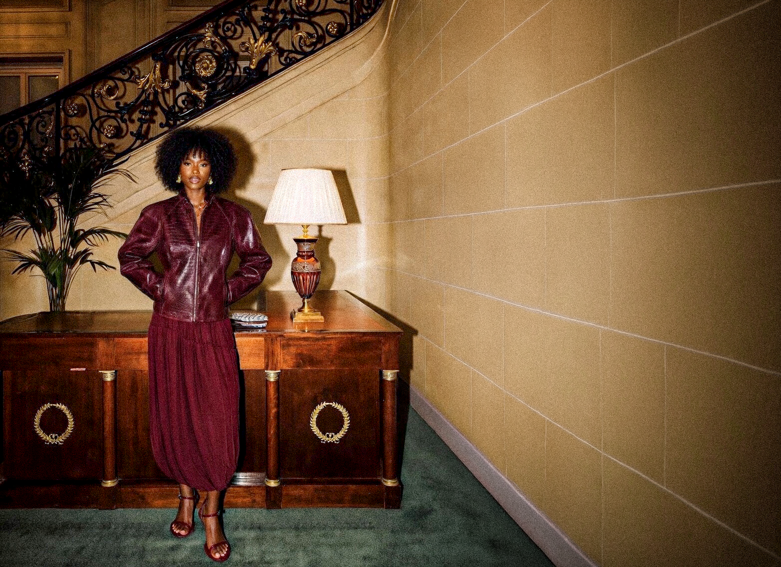 Woman in a maroon skirt and leather jacket by Adeptt standing in a room with a wooden desk and lamp.